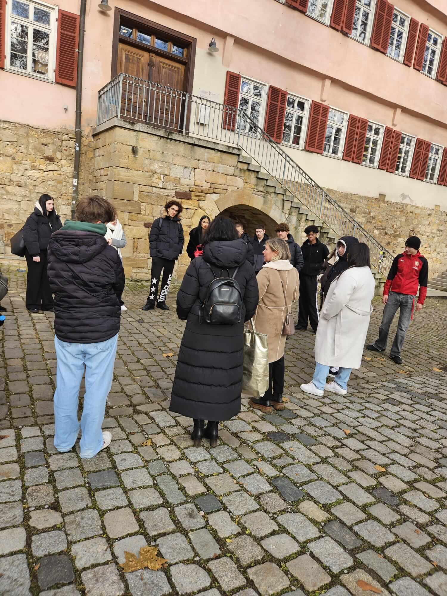 Eine Gruppe von Menschen steht auf einem gepflasterten Platz vor einem historischen Gebäude mit roten Fensterläden.