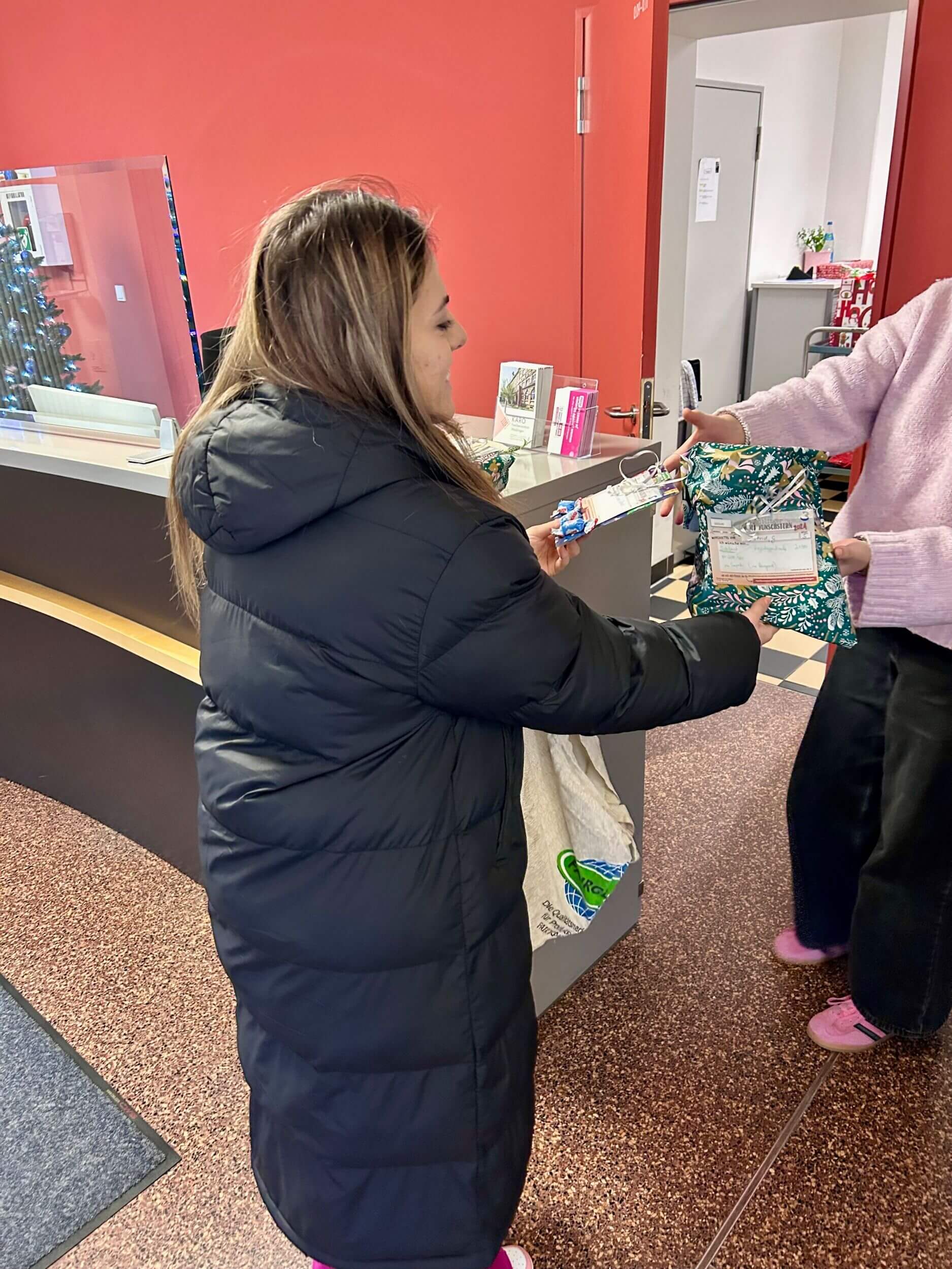 Eine Frau im schwarzen Mantel übergibt Geschenke in einem roten Empfangsbereich an eine andere Person.