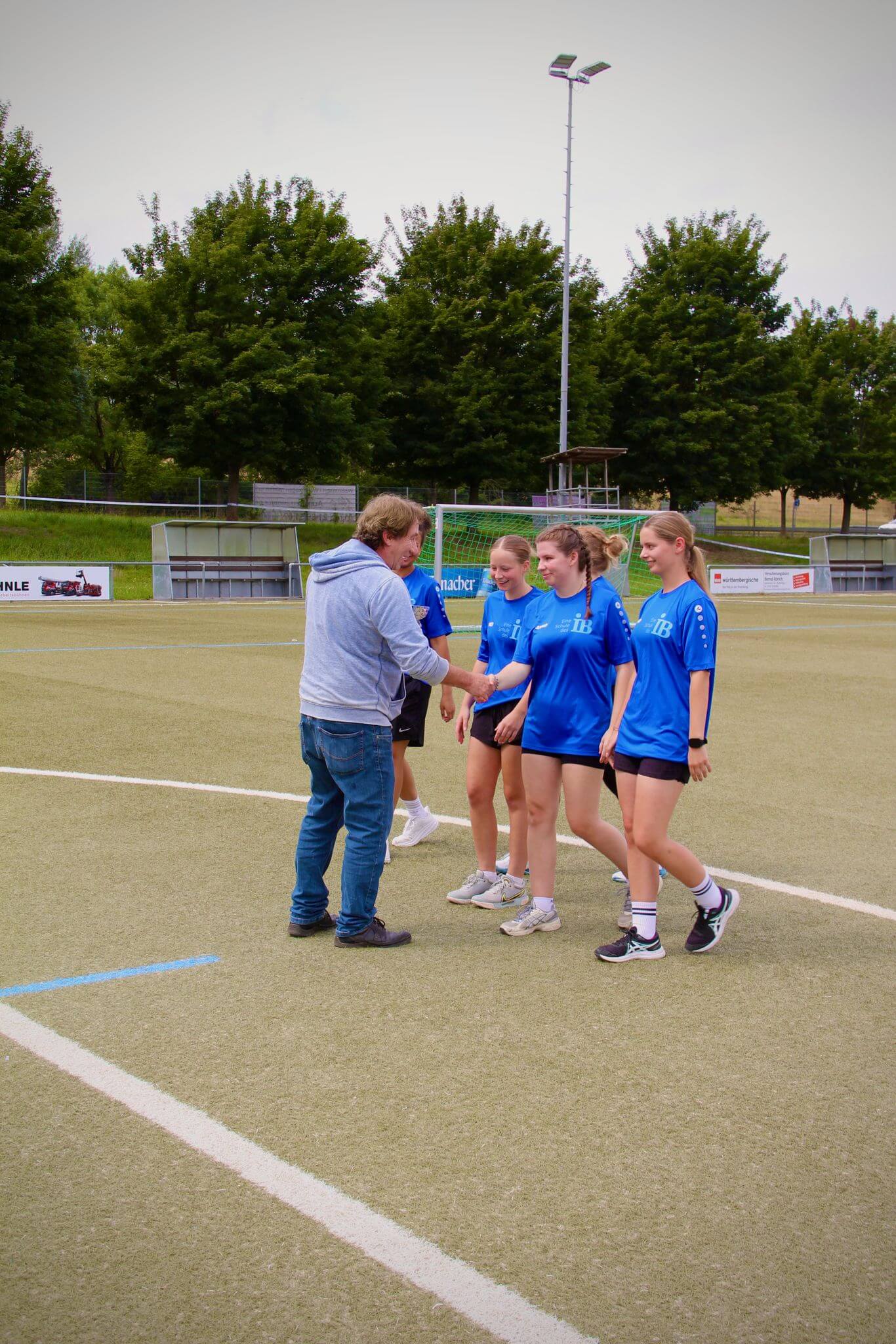 Eine Gruppe von jungen Frauen in blauen Trikots erhält auf einem Sportplatz Anweisungen von einem Mann.