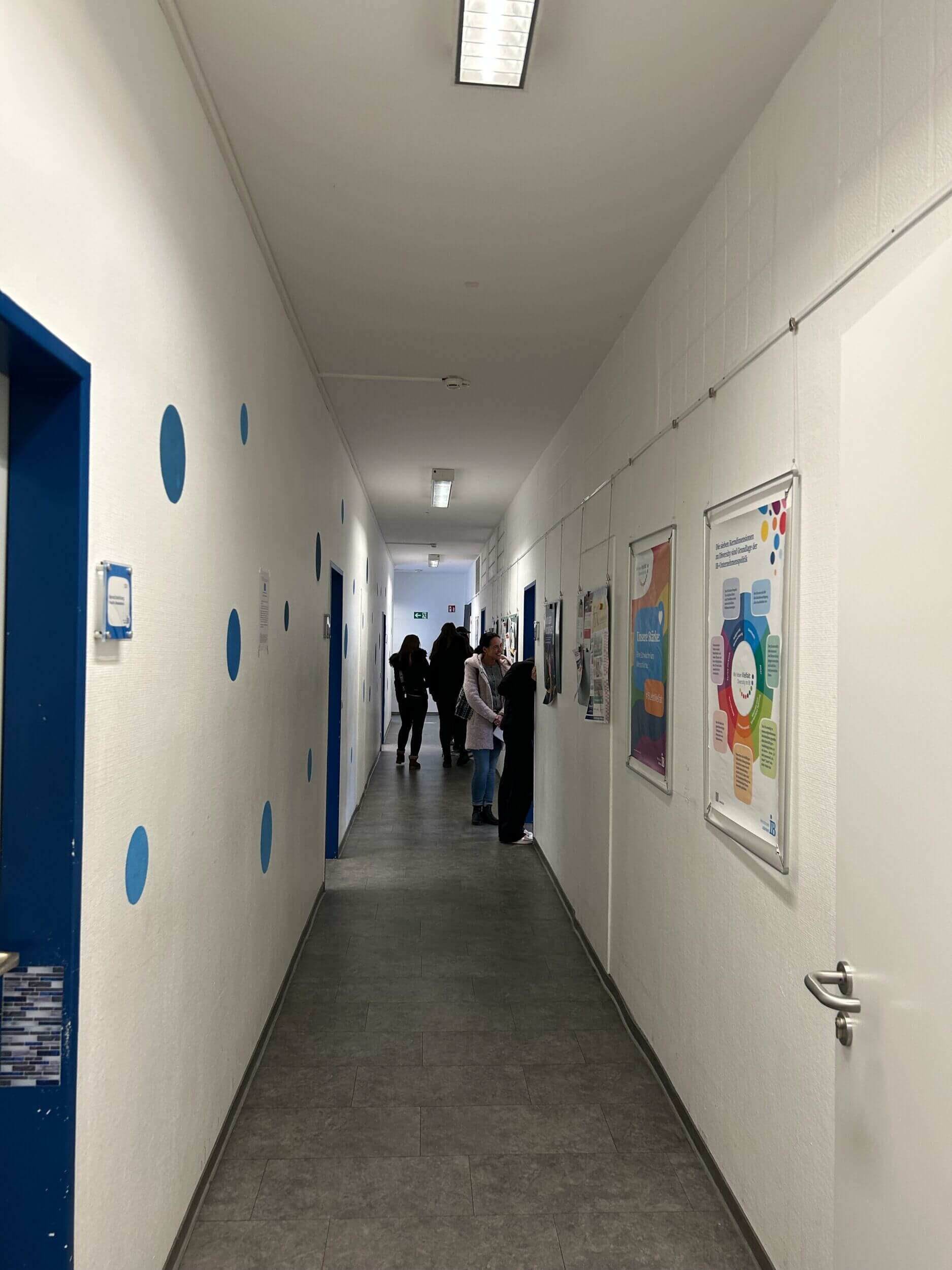 Ein langer Flur mit blauen Türen und Punkten zeigt Menschen, die Poster an der Wand betrachten.