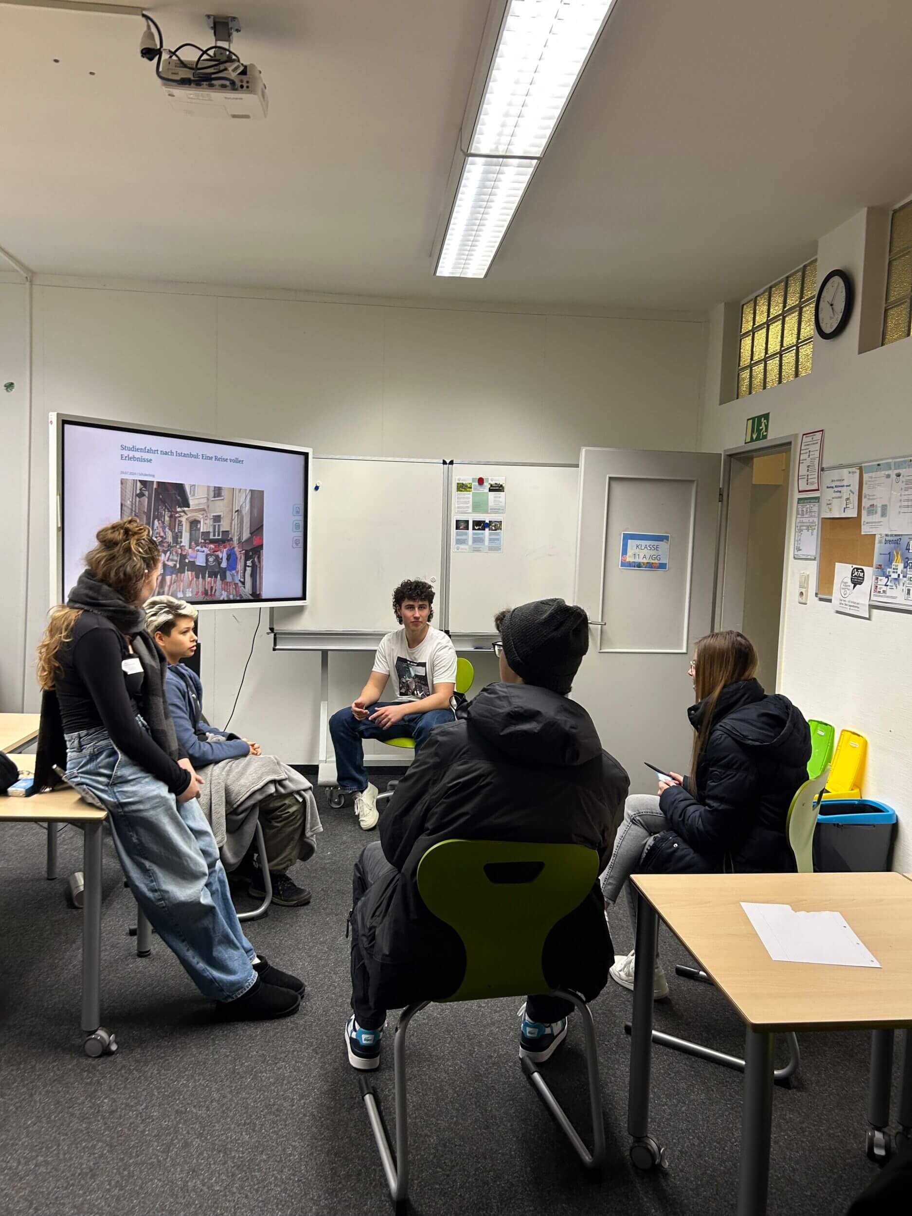 Eine Gruppe von Schülern diskutiert in einem Klassenzimmer über eine bevorstehende Studienfahrt, die auf einem Bildschirm angezeigt wird.