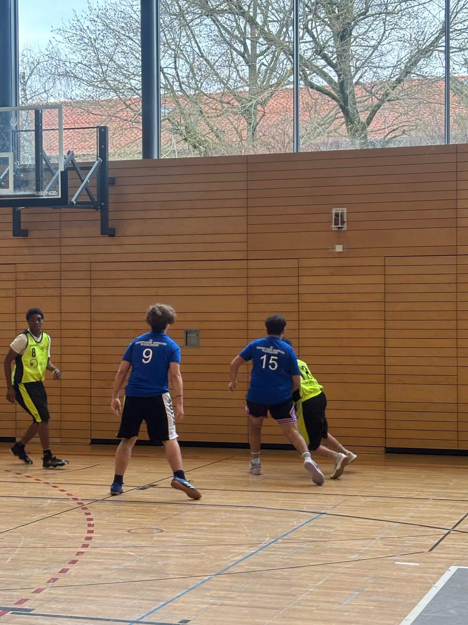 In einer Turnhalle spielen vier Männer in blauen und neonfarbenen Trikots Basketball vor großen Fenstern.