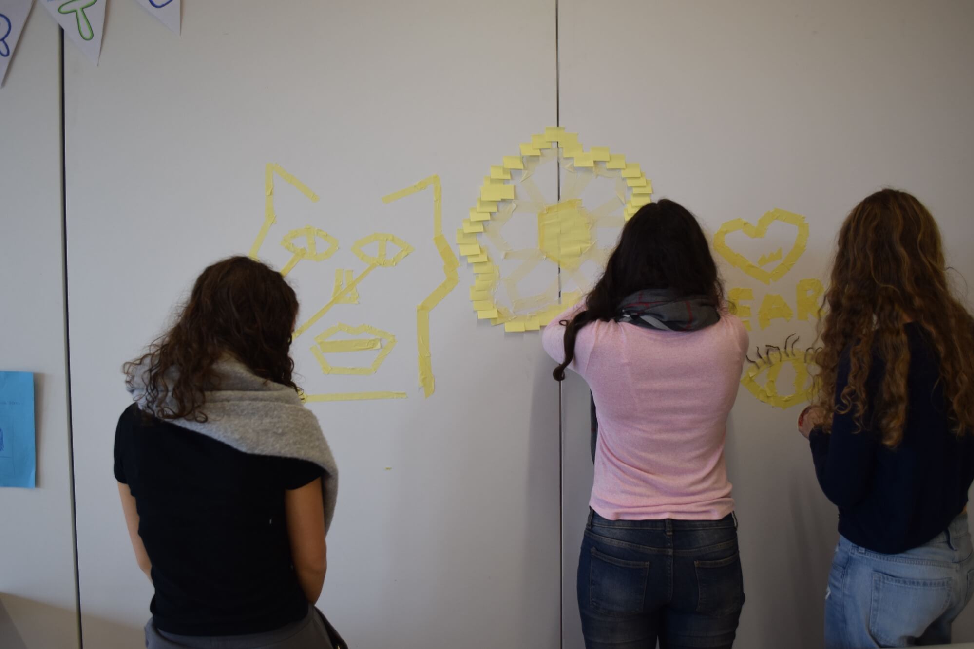 Drei Frauen kleben bunte Post-its an eine Wand, um kreative Kunstwerke in Form von Gesichtern und Herzen zu gestalten.