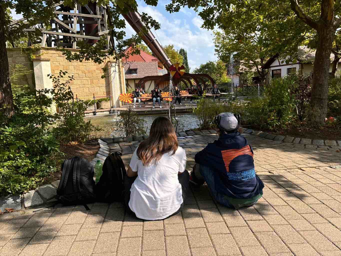 Zwei Personen sitzen im Schatten auf dem Bürgersteig und beobachten eine Wasserattraktion in einem Freizeitpark.
