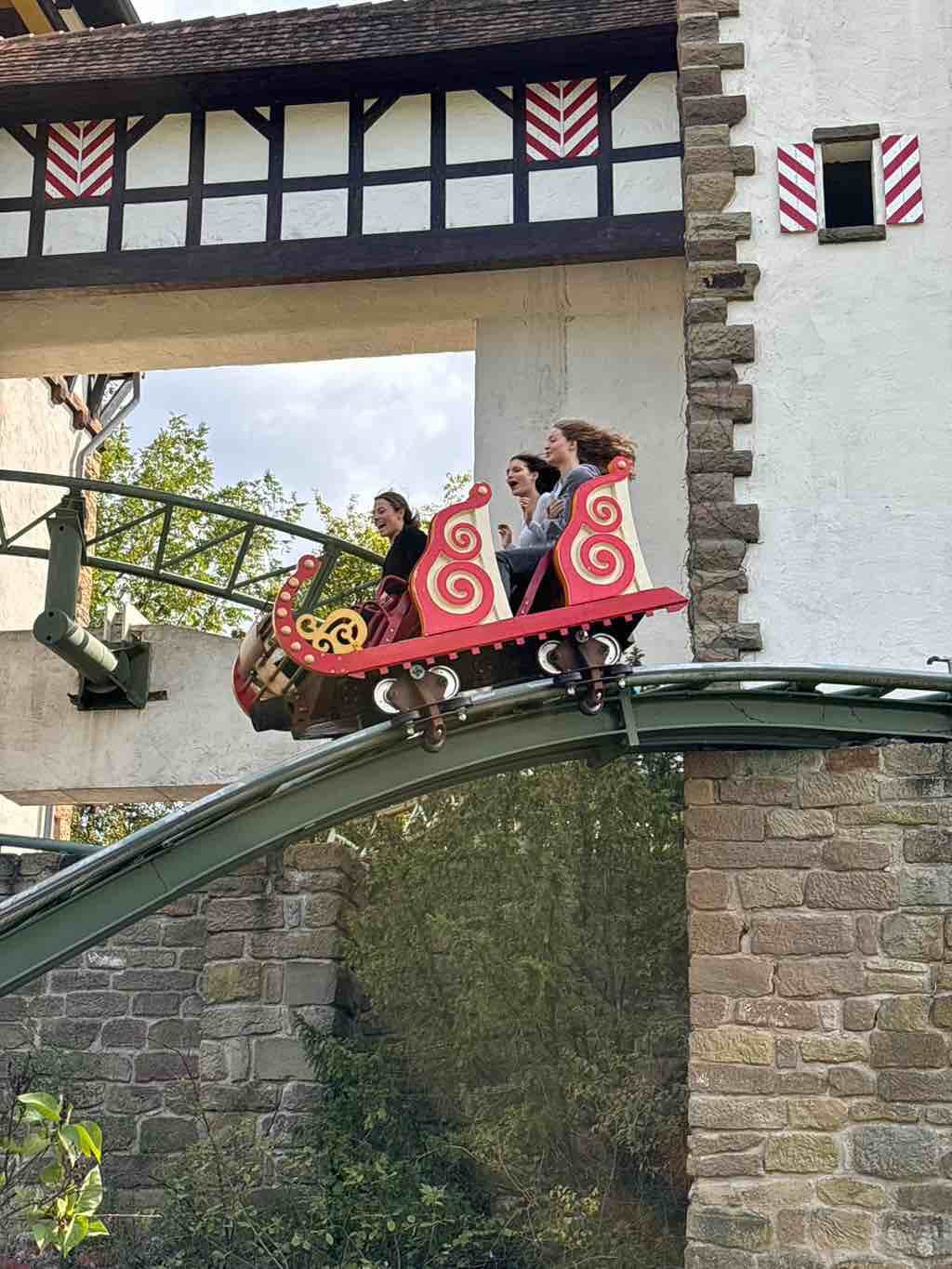 Drei Personen genießen eine aufregende Fahrt in einer roten Achterbahn, die durch eine mittelalterlich gestaltete Kulisse fährt.